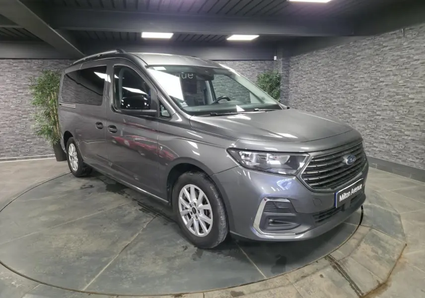 Vue 3/4 avant droit d'un Ford Transit Connect gris avec calandre noire et barres de toit argentées en intérieur showroom.