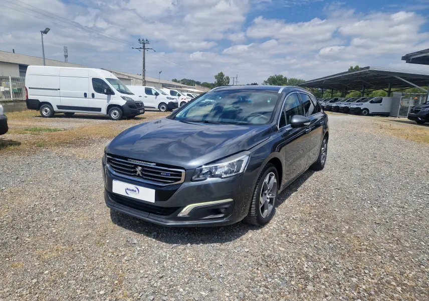 Peugeot 508 SW gris shark vue 3/4 avant droit sur parking gravillonné avec plusieurs utilitaires blancs en arrière-plan.