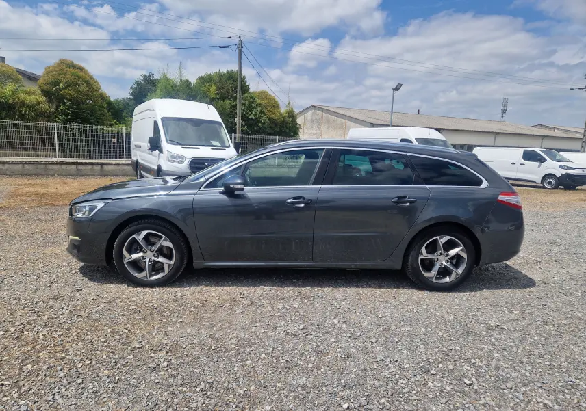 Profil côté gauche d'une Peugeot 508 SW gris shark 2015, break diesel avec jantes alliage 17 pouces sur sol gravillonné.