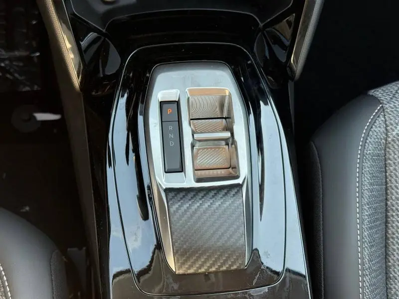 Vue plongeante sur la console centrale noire brillante de la Peugeot 208 Hybrid avec commandes de boîte automatique et insert carbone.