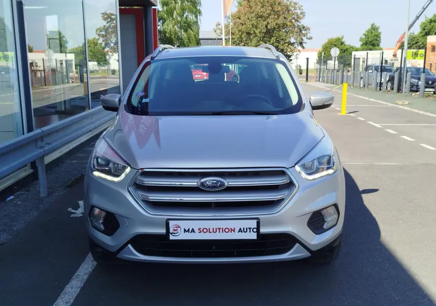 Vue frontale d'un Ford Kuga gris clair 2019 avec phares allumés et calandre chromée sur parking urbain.