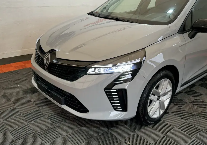 Vue 3/4 avant droit d'une Renault Clio gris clair 2025 avec phares LED allumés et jantes alliage.