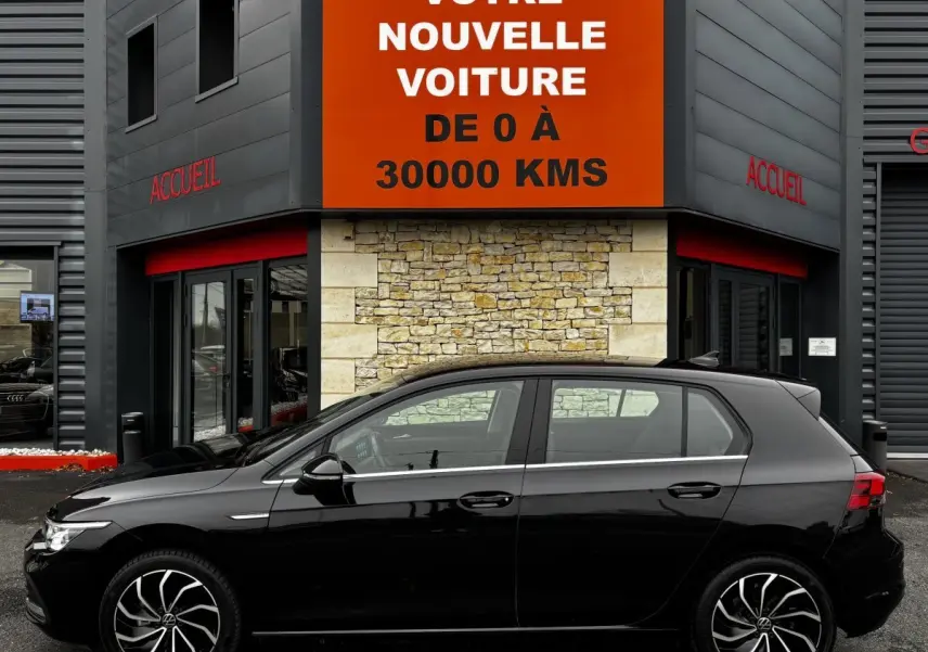 Profil côté gauche d'une Volkswagen Golf noire 2024 garée devant un bâtiment avec panneau orange "Votre nouvelle voiture".