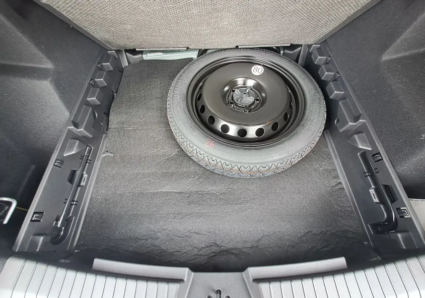 Vue plongeante sur la roue de secours noire dans le coffre du Dacia Bigster Hybrid 155 Journey + Pack Parking, intérieur gris.