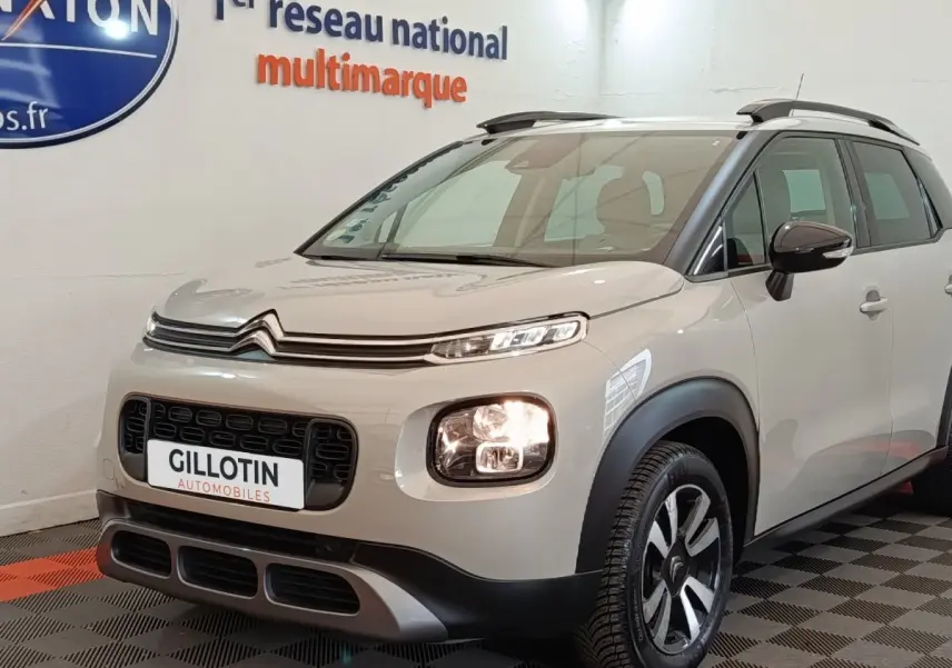 Vue 3/4 avant droite d'un Citroën C3 Aircross beige avec feux allumés et barres de toit noires en intérieur showroom.