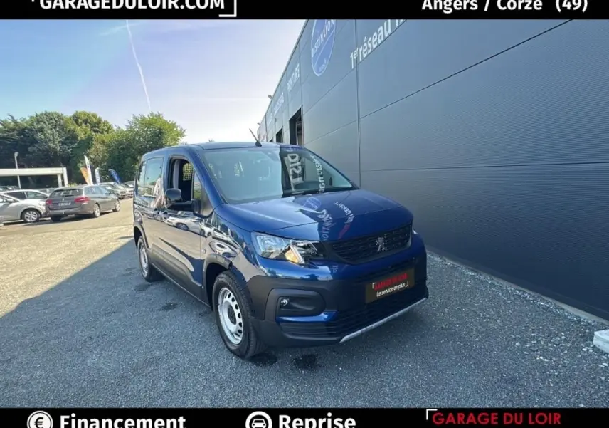 Peugeot Rifter bleu 2024 en 3/4 avant droit, avec calandre noire et jantes acier, stationné devant un bâtiment gris.