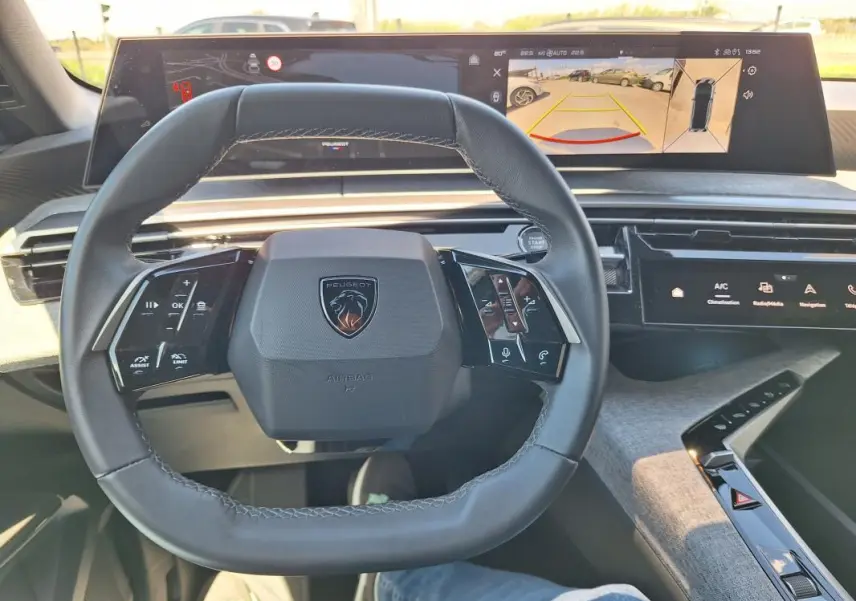 Vue intérieure centrée sur le volant cuir du Peugeot 3008 Hybrid 2025 avec cockpit numérique et écran d’aide au stationnement.