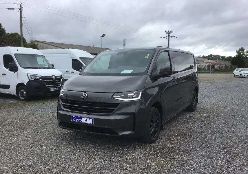 Vue 3/4 avant d'un Volkswagen Transporter T7 gris anthracite avec projecteurs LED Matrix et jantes noires sur parking gravel.
