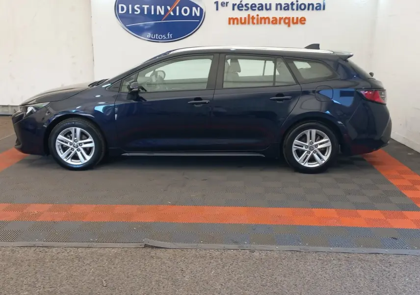 Vue latérale gauche d'une Toyota Corolla bleue hybride 2022 en version break, stationnée en intérieur showroom.