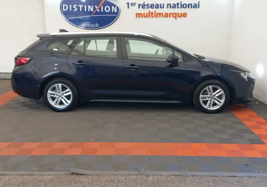 Profil côté gauche d'une Toyota Corolla break hybride bleu foncé, avec jantes alliage et toit blanc en intérieur showroom.