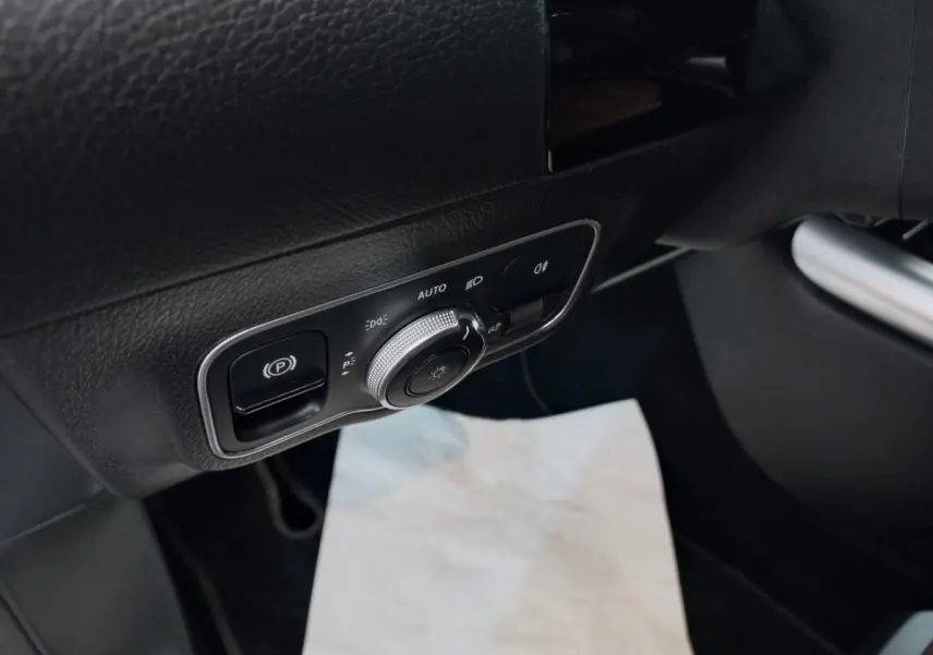 Gros plan sur le panneau de commande d’éclairage et frein à main électrique côté gauche intérieur d’une Mercedes GLA gris foncé.