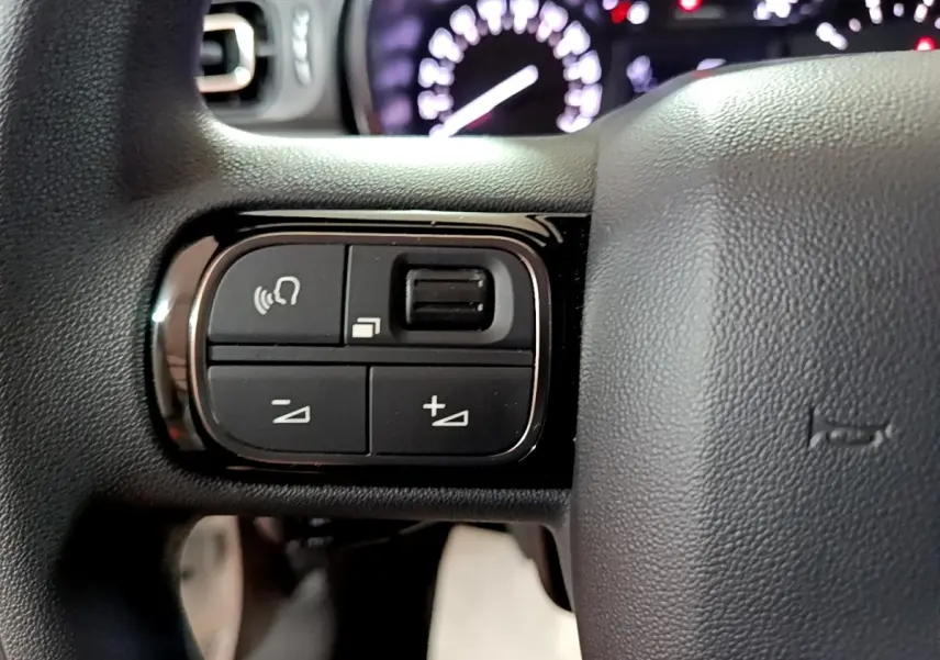 Gros plan sur les commandes du volant à gauche de la Citroën C3 blanche, tableau de bord flou en arrière-plan.