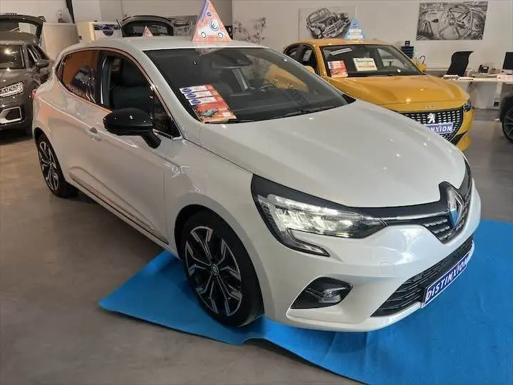 Renault Clio V blanc nacré métal vue 3/4 avant droit en showroom avec jantes alliage noires et toit noir.