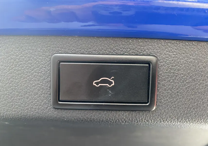 Bouton noir de déverrouillage du coffre sur fond de plastique texturé et partie de carrosserie bleu énergie d'une Skoda Superb Combi IV.