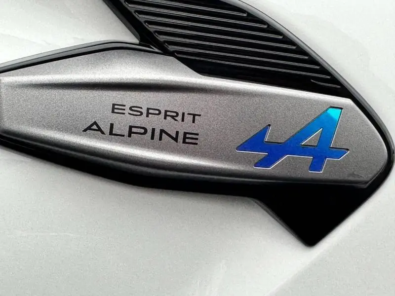 Détail du logo Esprit Alpine bleu sur fond gris métallisé du Renault Captur hybride 2025, vue rapprochée extérieure.