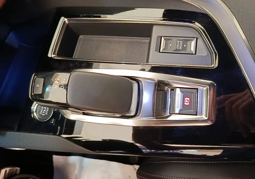 Gros plan sur la console centrale noire brillante du Peugeot 3008 Hybrid 225, avec levier de vitesse et bouton de frein à main électrique.