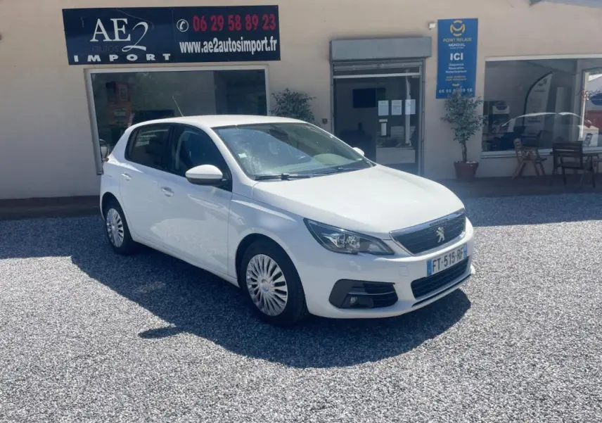 PEUGEOT 308 blanc vue 3/4 avant droit, garée devant un bâtiment avec panneaux publicitaires visibles.