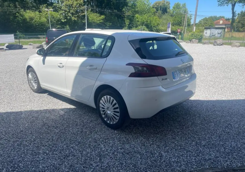 Peugeot 308 blanc vue 3/4 arrière côté gauche sur parking gravillonné, avec feux arrière foncés et jantes spécifiques.