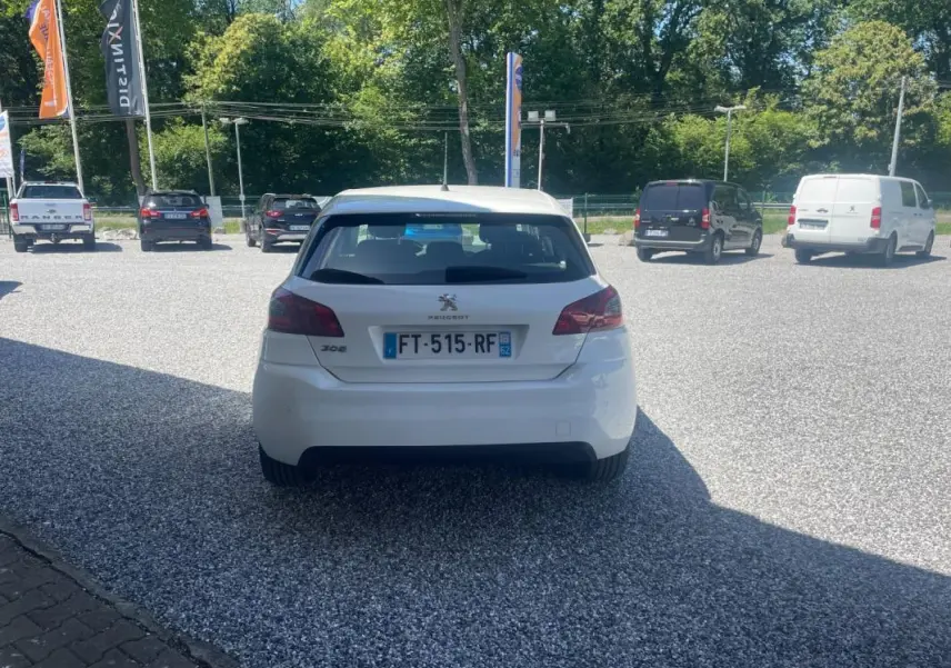 Vue arrière d'une Peugeot 308 blanche de 2020, stationnée sur un parking en gravier, avec plaques d'immatriculation visibles.