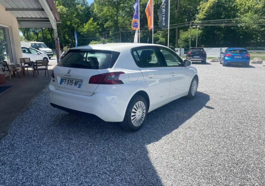 Peugeot 308 blanc vue 3/4 arrière droit sur parking gravillonné avec drapeaux publicitaires en arrière-plan.