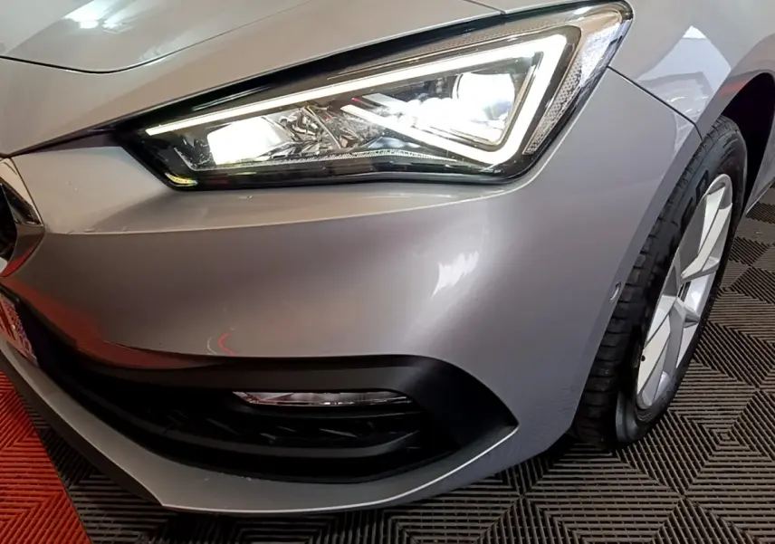 Gros plan sur le phare avant droit allumé d'une SEAT LEON gris foncé 2023 avec jante et pare-chocs noirs.