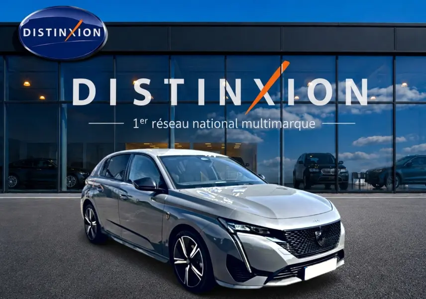 Peugeot 308 gris en 3/4 avant droit, avec calandre noire et jantes alliage distinctives, devant showroom Distinxion.