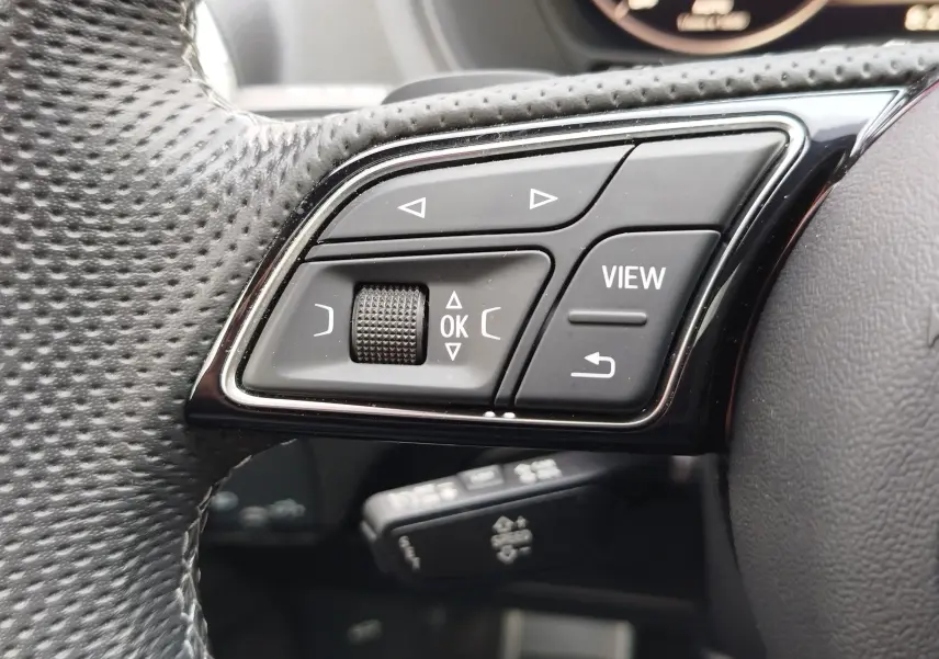 Gros plan sur les commandes gauche du volant noir perforé de l'Audi Q2 35 TFSI 2023, avec boutons de navigation et fonction View.