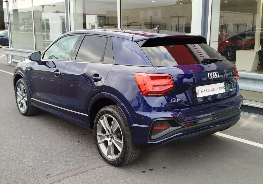 Audi Q2 bleu Navarre vue 3/4 arrière droit, avec jantes alliage et vitres teintées, stationnée devant un showroom.