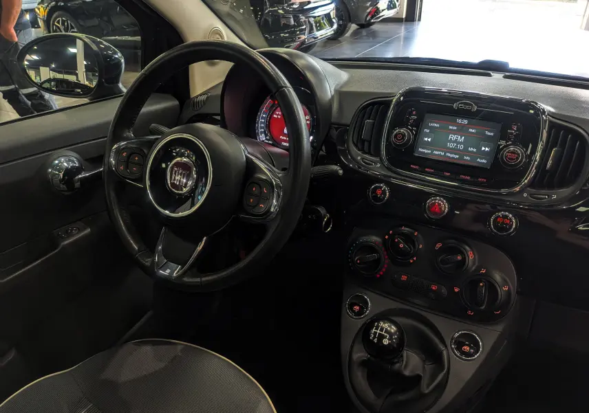 Vue intérieure côté conducteur de la Fiat 500 1.2 69 CH Lounge noire, avec volant multifonction et tableau de bord lumineux.