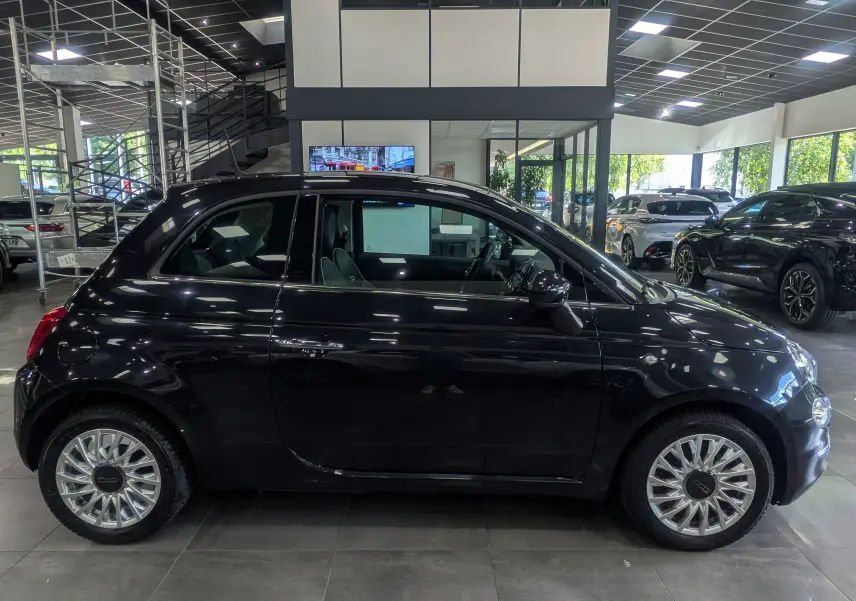 Profil côté gauche d'une FIAT 500 1.2 69 CH LOUNGE noire Crossover Black, en intérieur de showroom avec jantes argentées.