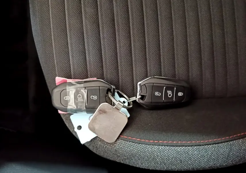 Gros plan sur deux clés noires posées sur un siège tissu gris foncé avec surpiqûres rouges d'une Peugeot 208 électrique blanche.