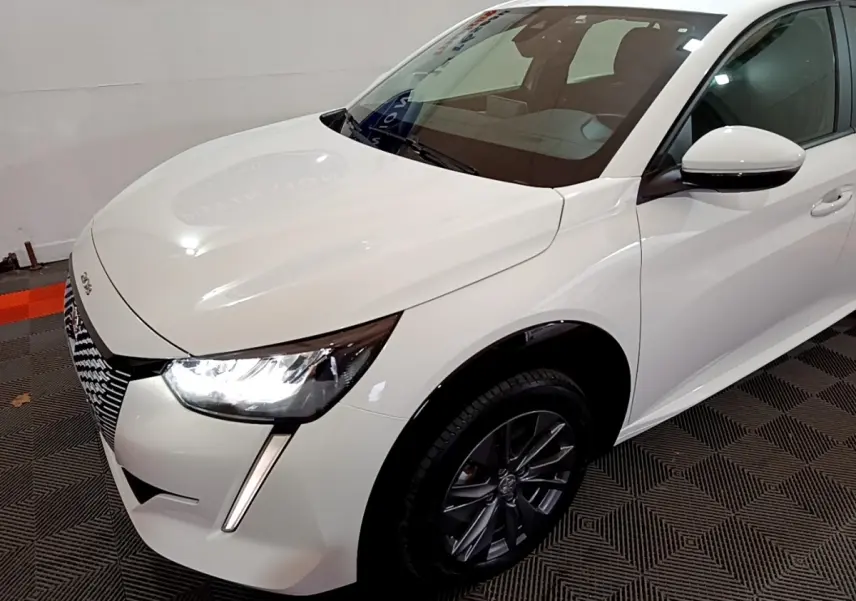 Vue 3/4 avant droit d'une Peugeot 208 électrique Active Business blanche avec phares LED allumés et jantes foncées.