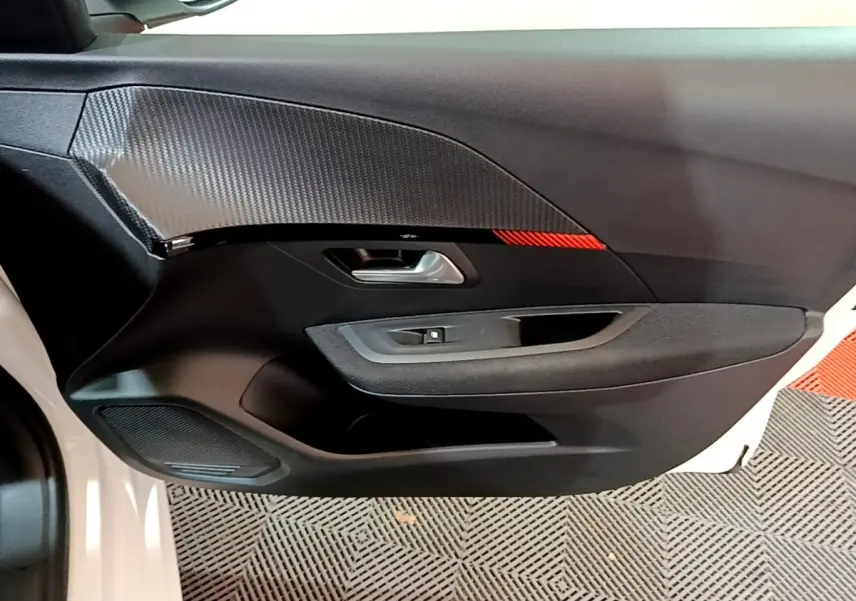 Détail de la porte avant gauche noire avec insert carbone et bande rouge d’une Peugeot 208 électrique blanche vue de l’intérieur.