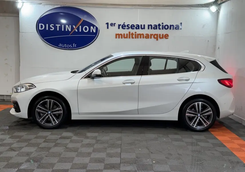 Profil droit d'une BMW Série 1 blanche 116i DKG7 Business Design garée en intérieur avec jantes alliage visibles.