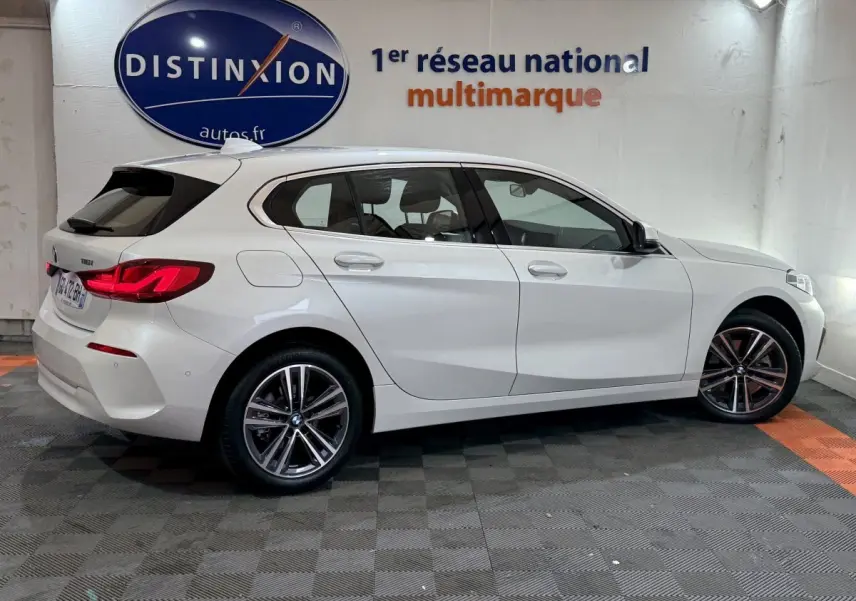 BMW Série 1 116i Business Design blanche vue de profil côté droit dans un showroom avec logo Distinxion en arrière-plan.
