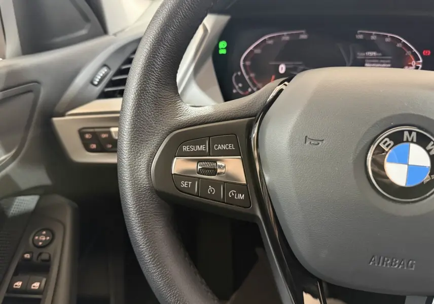 Gros plan sur le volant cuir noir de la BMW Série 1 116i 2023 avec commandes de régulateur et logo central visible.