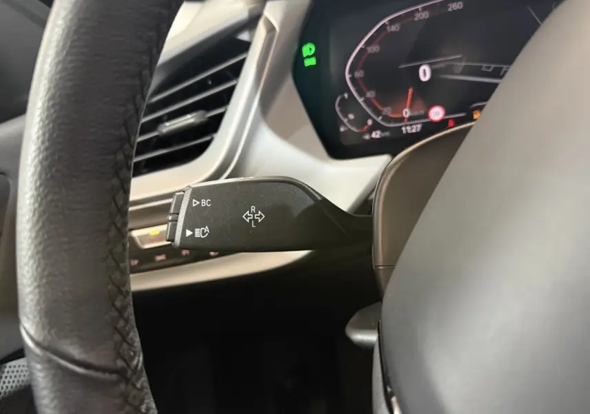 Gros plan sur la manette de clignotants noire à gauche du volant cuir dans le tableau de bord numérique d'une BMW Série 1 blanche 2023.