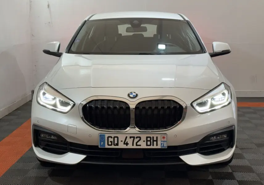 Vue frontale d'une BMW Série 1 116i blanche avec phares LED allumés et calandre noire caractéristique.