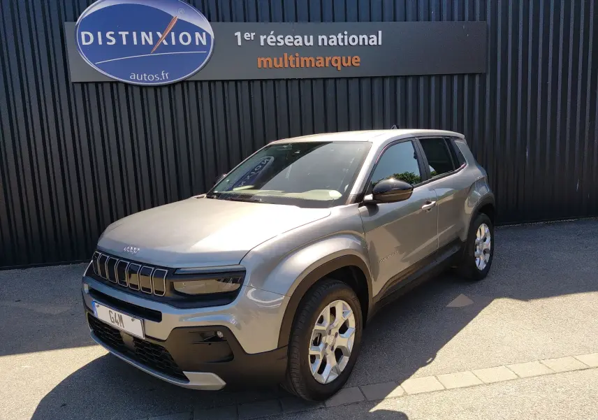 Jeep Avenger eHYBRID gris en 3/4 avant droit, avec calandre distinctive et jantes argentées, stationné devant un mur noir.