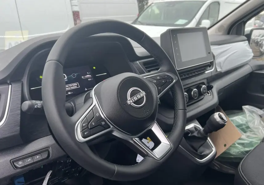 Vue intérieure côté conducteur du Nissan Primastar blanc 2025, montrant les trois sièges avant en tissu noir et le tableau de bord.