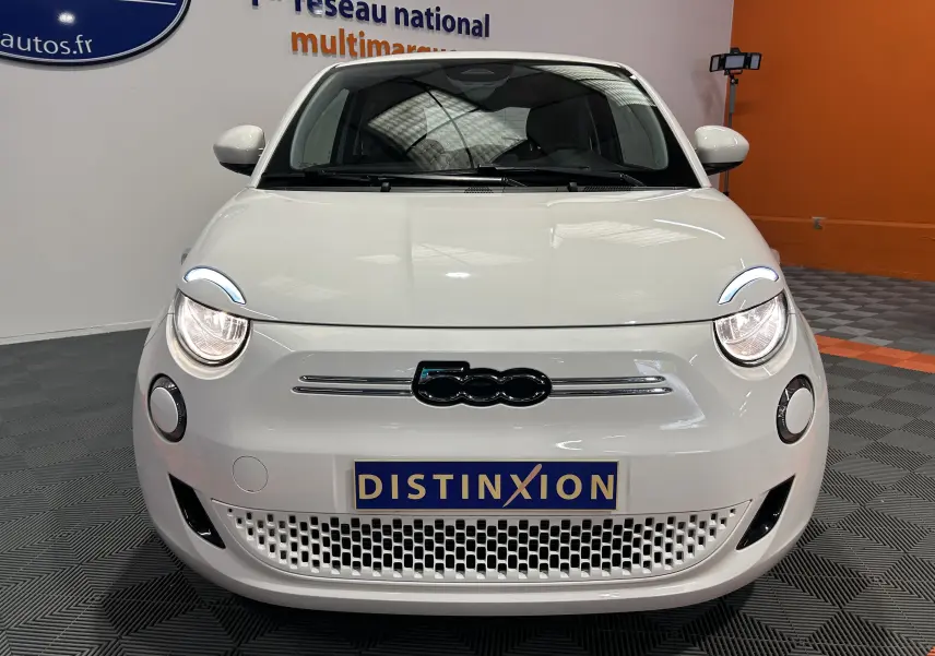Vue frontale d'une FIAT 500 électrique blanche 2023 avec phares allumés et logo 500 noir sur fond blanc.