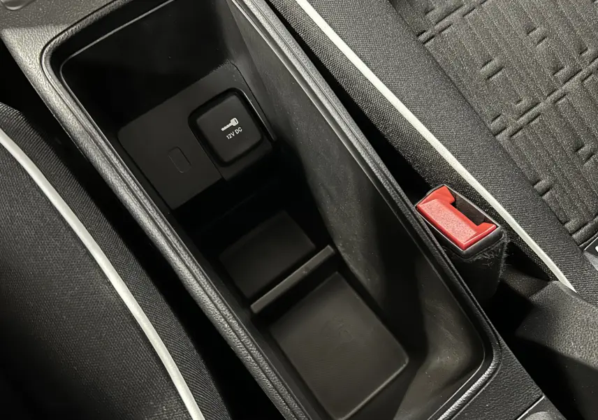 Gros plan sur la console centrale noire avec prise 12V et bouton de verrouillage, siège tissu gris avec surpiqûres blanches.