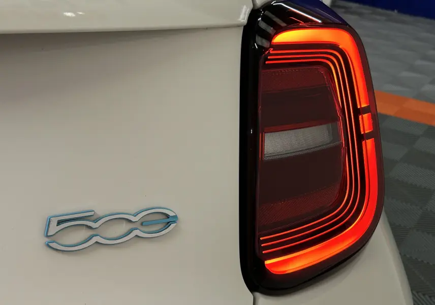 Gros plan sur le feu arrière droit allumé et le logo 500 bleu sur la carrosserie blanche d’une Fiat 500 électrique.