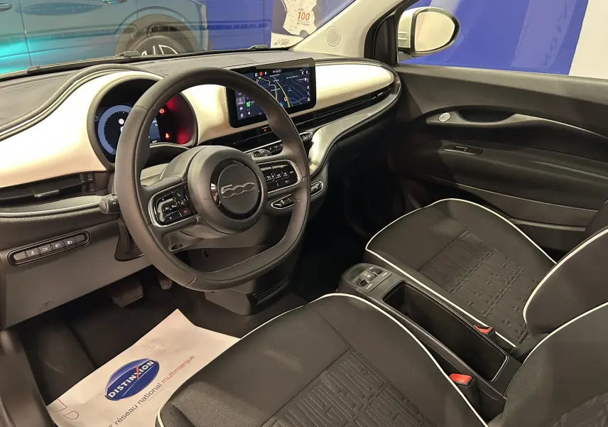 Vue intérieure côté conducteur de la Fiat 500 électrique 2023, tableau de bord blanc et sièges noirs avec surpiqûres blanches.