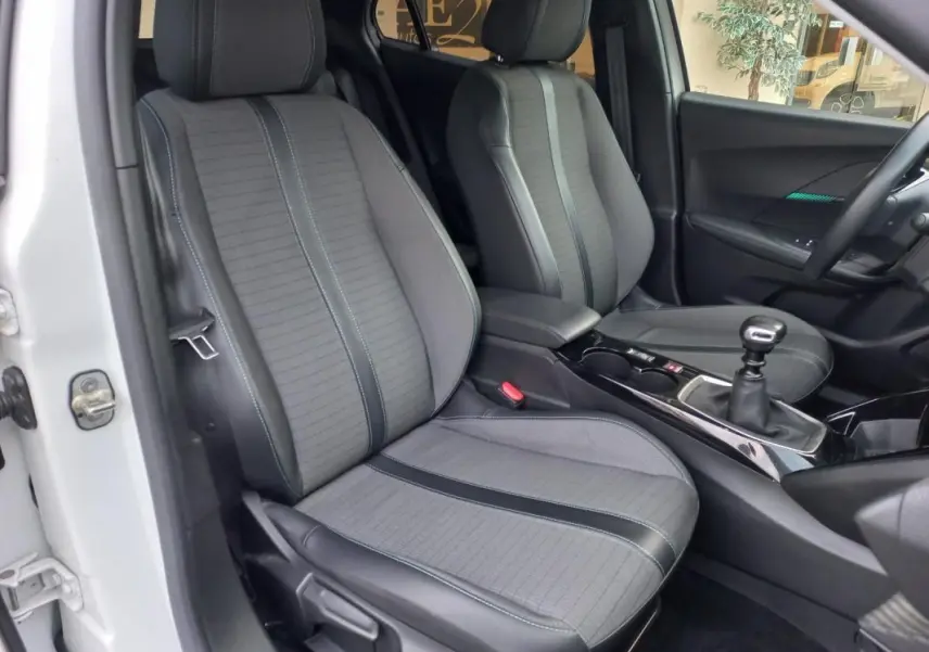 Vue intérieure côté conducteur de la Peugeot 2008 blanche, montrant les sièges gris et la boîte manuelle.