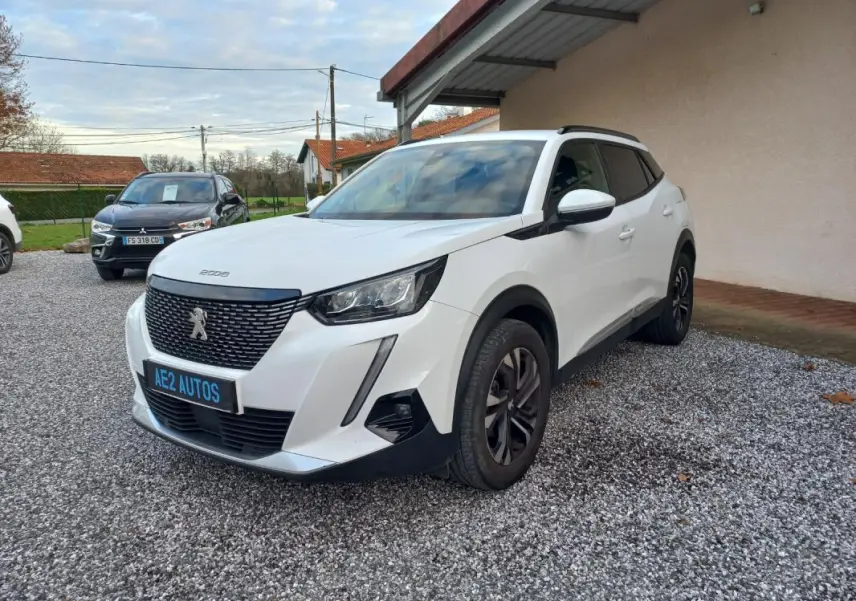 Peugeot 2008 blanc en 3/4 avant droit, avec calandre noire et jantes alliage foncé, stationné sur gravier.