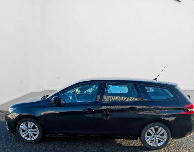 Vue latérale droite d'une Peugeot 308 SW noire stationnée devant un mur blanc avec logos en arrière-plan.