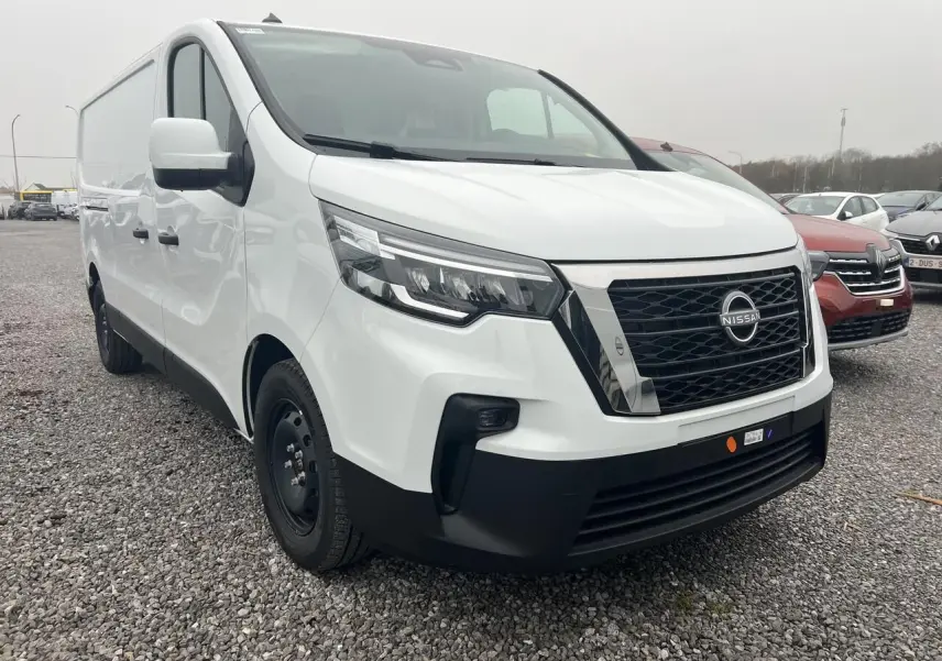 Vue 3/4 avant droit d’un utilitaire Nissan Primastar blanc avec calandre noire et logo Nissan visible, sur parking extérieur.