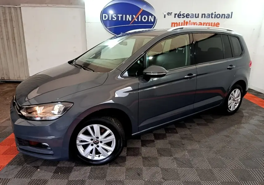 Vue 3/4 avant gauche d'un Volkswagen Touran gris foncé garé en intérieur avec jantes alliage et phares allumés.