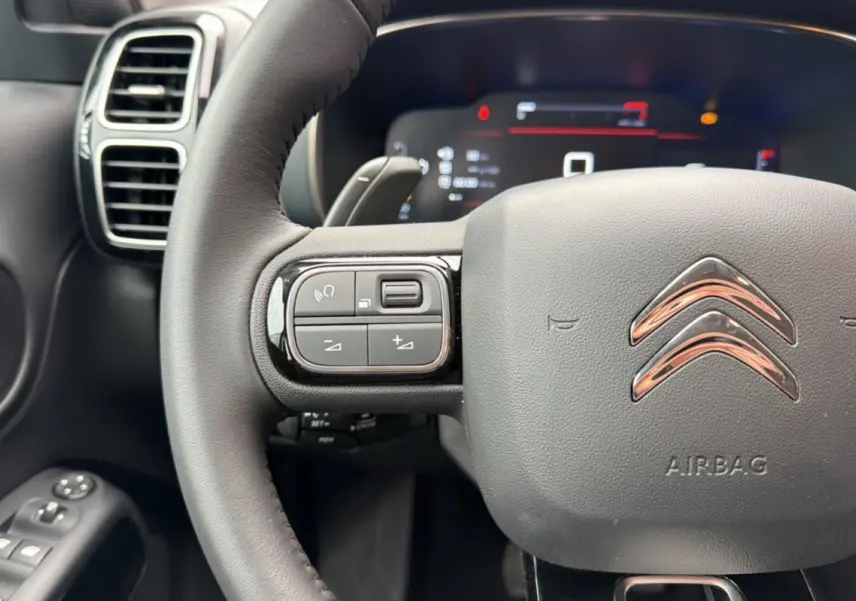 Gros plan sur le volant cuir multifonctions noir du Citroën C5 Aircross 2025 avec commandes intégrées et tableau de bord numérique flou.